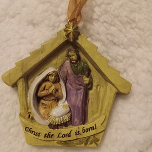 May include: A Christmas ornament depicting the nativity scene. The ornament shows Mary, Joseph, and baby Jesus in a stable. The text "Christ the Lord is born!" is on a banner. The ornament is made of light yellow material and has a gold ribbon for hanging.
