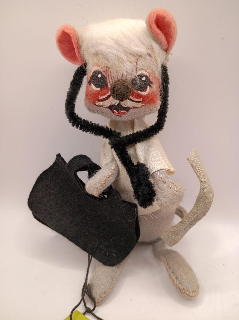 May include: A vintage stuffed mouse doll dressed as a doctor, wearing a black stethoscope and holding a black bag. The mouse has a white head with pink ears and a painted face with rosy cheeks.