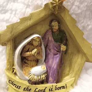May include: A Christmas ornament showing the Nativity scene. The ornament features Mary, Joseph, and baby Jesus in a manger, with the text "Christ the Lord is born!" in a banner below. The figures are in a stable-like structure.