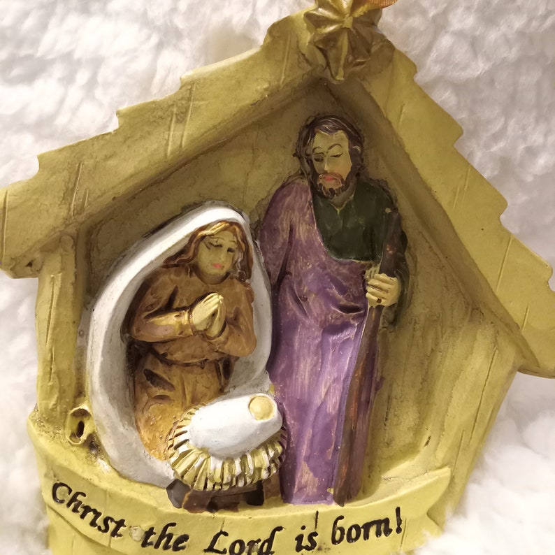 May include: A Christmas ornament showing the Nativity scene. The ornament features Mary, Joseph, and baby Jesus in a manger, with the text "Christ the Lord is born!" in a banner below. The figures are in a stable-like structure.