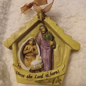 May include: A beige Christmas ornament depicting the Nativity scene. The ornament features Mary, Joseph, and baby Jesus in a manger, with the text "Christ the Lord is born!" on a banner. A gold ribbon is attached for hanging.