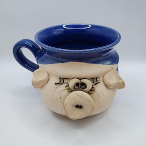 May include: A ceramic mug with a pig face design. The mug has a blue interior and handle, with a cream-colored body featuring a pig snout, ears, and eyes. The mug is handmade and has a whimsical, rustic aesthetic.