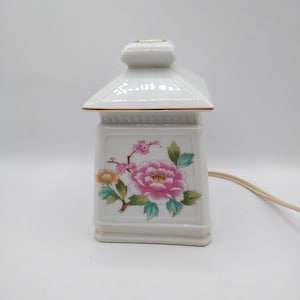 May include: White ceramic lantern-shaped lamp with a floral design. The lamp features a pink peony and other flowers with green leaves. Gold trim accents the edges. The lamp has a square base and a small, square top.