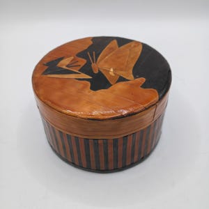 May include: A round wooden box with a lid featuring a butterfly and leaf design. The box is brown with black vertical stripes around the sides. The lid has a butterfly and leaf design in shades of brown and black.