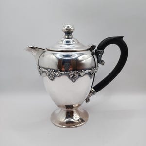 May include: A silver-colored teapot with a black handle and a decorative floral band around the middle. The teapot has a lid with a small knob and a spout for pouring. The base is a pedestal design.