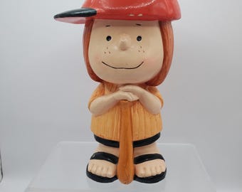May include: A ceramic figurine of a girl wearing a red baseball cap, a yellow and orange striped shirt, and black sandals. She is holding a wooden baseball bat and has a smiling face.