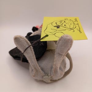 May include: A handmade gray and black stuffed animal mouse with a pink flower and a yellow tag. The tag features a drawing of a girl and the words "Annada Dolls". The mouse has a long, curved tail.