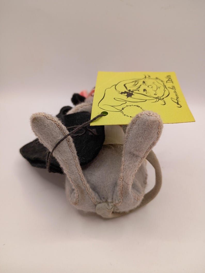 May include: A handmade gray and black stuffed animal mouse with a pink flower and a yellow tag. The tag features a drawing of a girl and the words "Annada Dolls". The mouse has a long, curved tail.