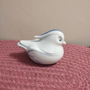 May include: A white ceramic bird figurine with blue accents. The bird has a long, thin beak and a blue stripe on its head. The bird is sitting on a pink knitted surface.