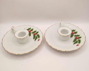 May include: A pair of white porcelain candle holders with a scalloped edge and gold trim. Each holder features a central cup with a small handle and a festive green holly leaf and red berry pattern painted on the side of the circular base.