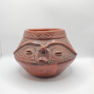 May include: A terracotta-colored ceramic pot with a stylized face design. The pot features etched details around the eyes, mouth, and upper rim. The vessel has a rounded body and a wide opening, showcasing a unique, handcrafted aesthetic.