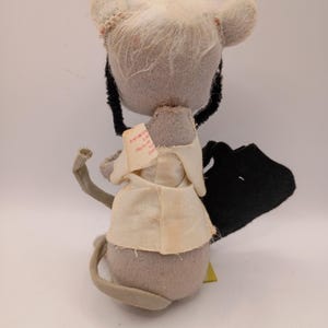 May include: A vintage stuffed mouse toy with a gray body, white fur, and a cream-colored outfit. The mouse has black whiskers and a long gray tail. A small tag is attached to the outfit.
