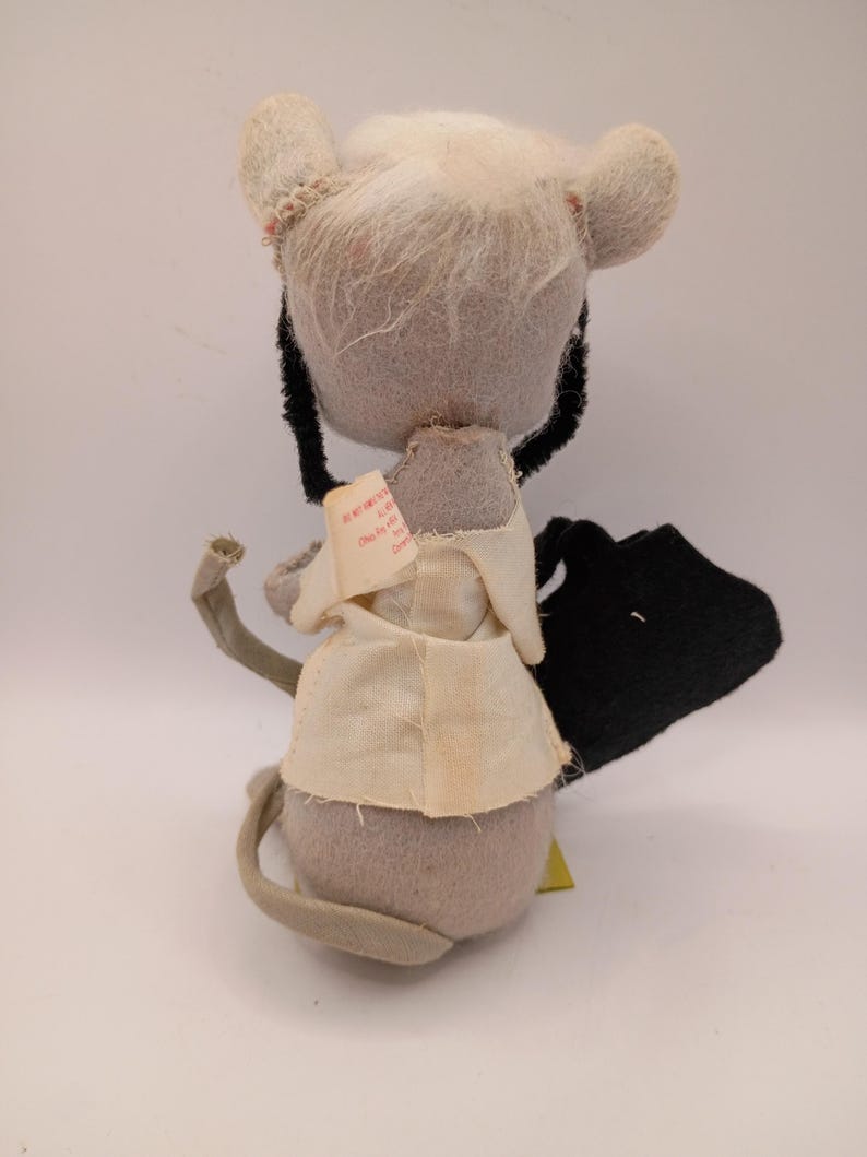 May include: A vintage stuffed mouse toy with a gray body, white fur, and a cream-colored outfit. The mouse has black whiskers and a long gray tail. A small tag is attached to the outfit.