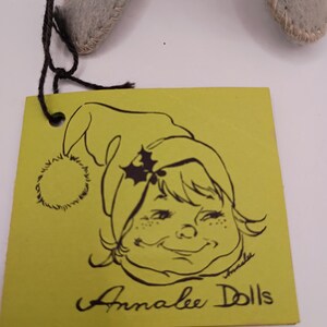 May include: A yellow tag with a black illustration of a smiling girl wearing a green hat with holly. The tag reads "Annalee Dolls" in black script. The tag is attached to a gray felt doll.