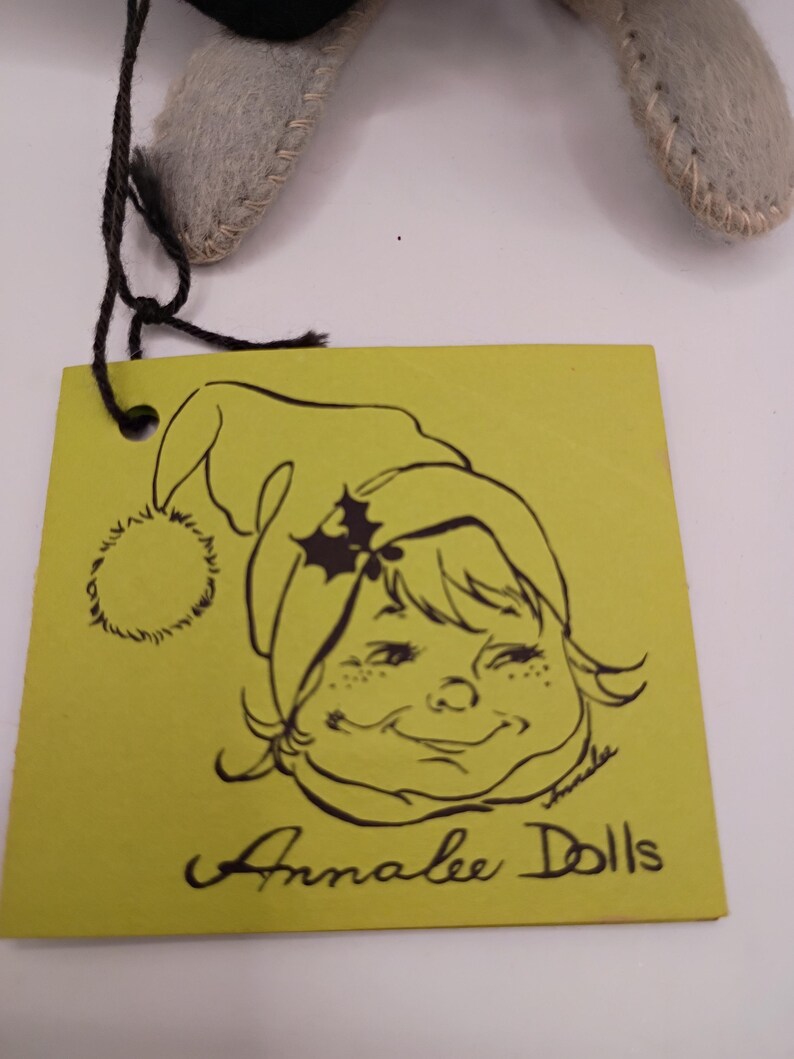 May include: A yellow tag with a black illustration of a smiling girl wearing a green hat with holly. The tag reads "Annalee Dolls" in black script. The tag is attached to a gray felt doll.