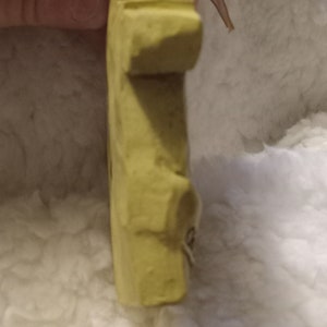 May include: A light yellow, rectangular, carved object with a rounded top and a small, attached string. The object has a rough texture and appears to be handmade. The background is a soft, white, fluffy material.