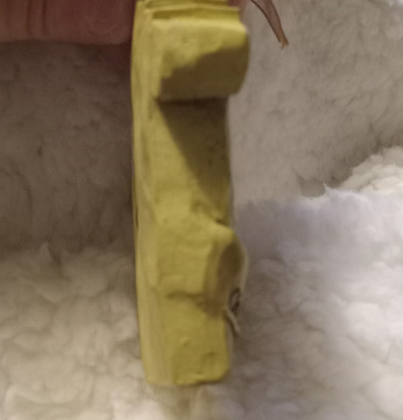 May include: A light yellow, rectangular, carved object with a rounded top and a small, attached string. The object has a rough texture and appears to be handmade. The background is a soft, white, fluffy material.