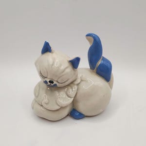 May include: A ceramic figurine of a sleeping cat. The cat is primarily white with blue accents on the ears, tail, and nose. The cat is curled up in a relaxed pose, with its eyes closed.
