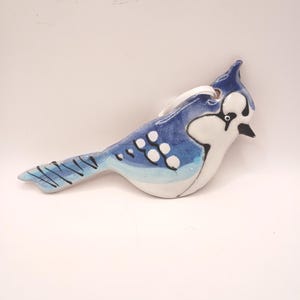 May include: A ceramic blue jay ornament with a white belly and accents. The bird has a detailed design with black lines and white spots. It has a loop for hanging.
