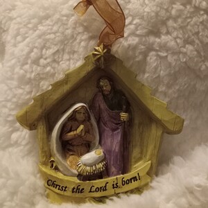 May include: A Christmas ornament showing the Nativity scene. The ornament features Mary, Joseph, and baby Jesus in a manger, with the text "Christ the Lord is born!" on a banner. The ornament is made of a beige material and has a gold ribbon for hanging.