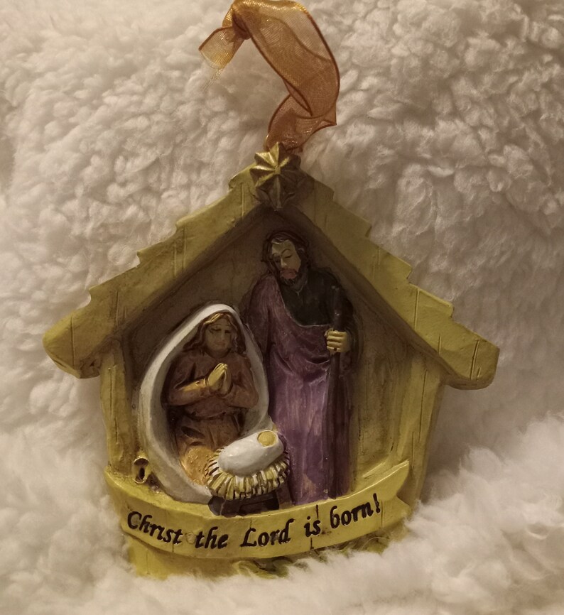 May include: A Christmas ornament showing the Nativity scene. The ornament features Mary, Joseph, and baby Jesus in a manger, with the text "Christ the Lord is born!" on a banner. The ornament is made of a beige material and has a gold ribbon for hanging.