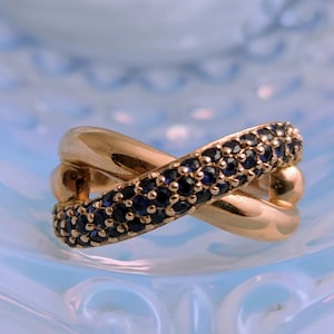 May include: A gold-colored ring with a crisscross design. One band is encrusted with small, dark blue gemstones. The ring is set against a light blue, textured background, creating a contrast between the warm gold and cool blue.