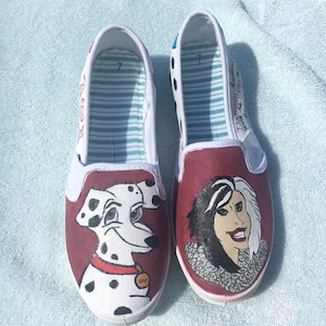 Può includere: Paio di scarpe slip-on bianche, taglia 7, con disegni dipinti a mano. Una scarpa raffigura un cane dalmata, l'altra Crudelia De Mon. Le scarpe hanno puntali bordeaux e fodere interne a righe. Lo sfondo è azzurro.