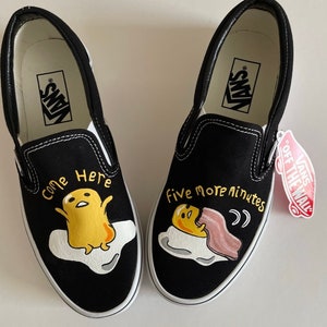 May include: Pair of black slip-on sneakers with white soles and trim. Each shoe features a cartoon egg character on a white cloud. One shoe says "Come Here", the other says "Five More Minutes". A Vans tag is attached.