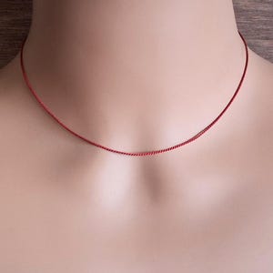 May include: A thin, vibrant red necklace chain resting against the neck. The chain features a delicate, twisted rope-like texture that creates a subtle shimmer. This minimalist piece of jewelry sits close to the base of the throat in a simple, circular design.