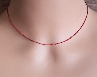 May include: A thin, vibrant red necklace chain resting against the neck. The chain features a delicate, twisted rope-like texture that creates a subtle shimmer. This minimalist piece of jewelry sits close to the base of the throat in a simple, circular design.