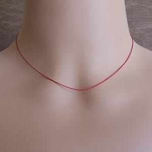 May include: A thin, vibrant red cord necklace resting against the neck. The necklace consists of a single, delicate strand of red thread that forms a simple circular shape, providing a minimalist and understated appearance around the collarbone area.