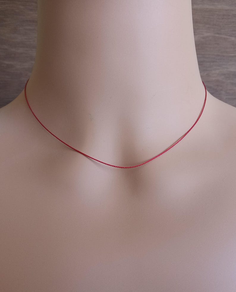 May include: A thin, vibrant red cord necklace that rests against the neck. The necklace consists of a single, smooth, and slender strand of red material, forming a simple circular shape that sits close to the base of the neck without any pendants.