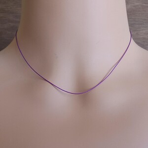 May include: A thin, vibrant purple cord necklace that rests against the neck. The necklace consists of a single, smooth, and slender strand of purple material, forming a simple and minimalist circular shape around the base of the neck.