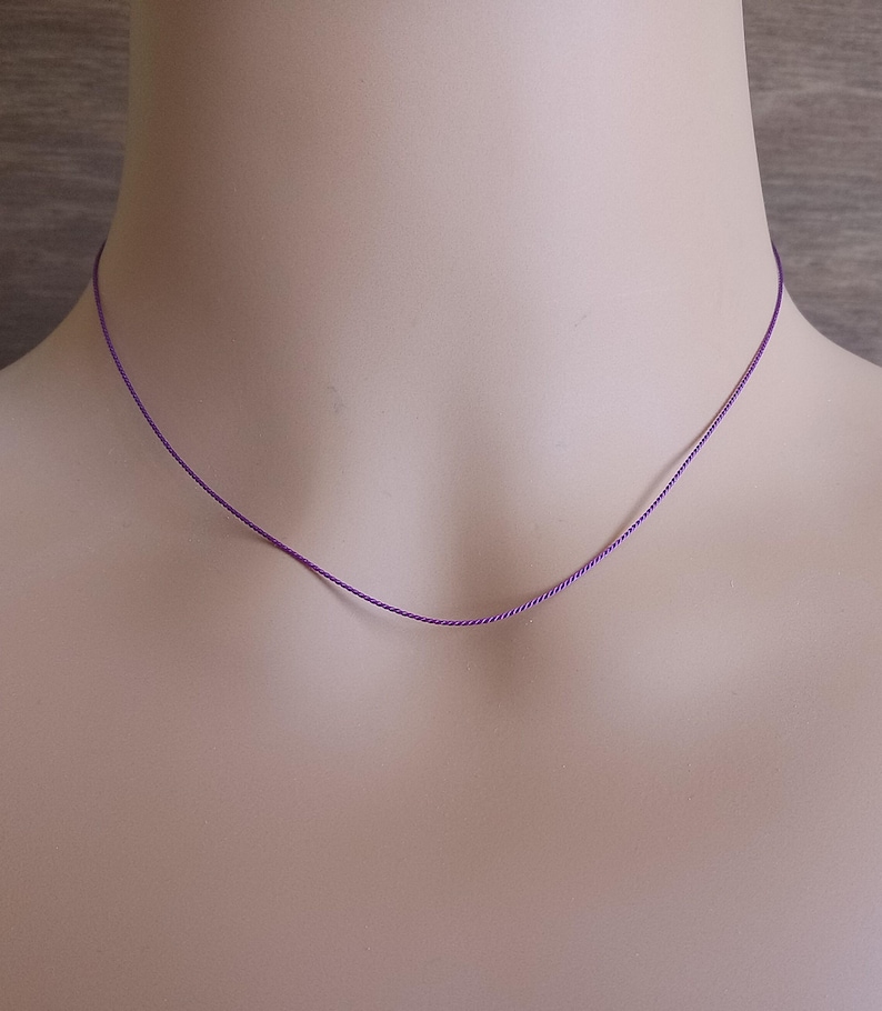 May include: A thin, vibrant purple cord necklace that rests against the neck. The necklace consists of a single, smooth, and slender strand of purple material, forming a simple and minimalist circular shape around the base of the neck.