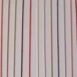 May include: A collection of fifteen vertical, thin, twisted cords arranged in parallel. The cords feature a variety of solid colors including green, purple, orange, yellow, black, red, white, light blue, dark blue, pink, brown, and gold.
