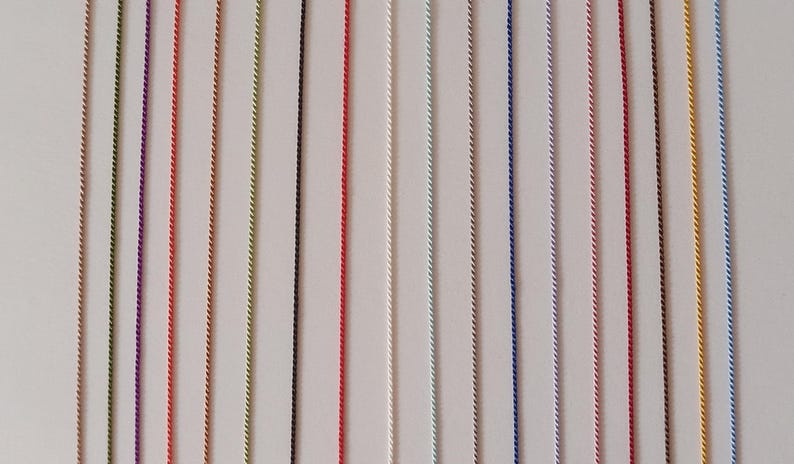 May include: A collection of fifteen vertical, thin, twisted cords arranged in parallel. The cords feature a variety of solid colors including green, purple, orange, yellow, black, red, white, light blue, dark blue, pink, brown, and gold.