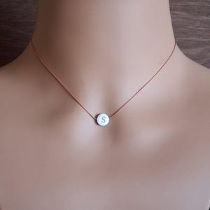 May include: A delicate necklace featuring a small, round pendant with the letter "S" in the center. The pendant is suspended from a thin, brown cord. The necklace is displayed on a mannequin.