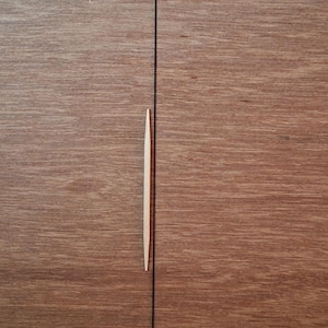 May include: A single, light-colored wooden toothpick with pointed ends positioned vertically over a dark brown wood grain surface that features a distinct, straight vertical seam running through the center of the frame.