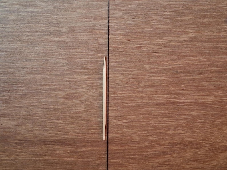 May include: A single, light-colored wooden toothpick with pointed ends positioned vertically over a dark brown wood grain surface that features a distinct, straight vertical seam running through the center of the frame.