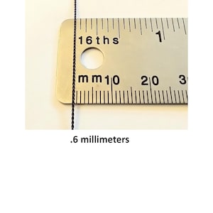 May include: A thin black twisted cord positioned vertically against a metal ruler. The ruler displays markings for 16ths of an inch and millimeters. The text .6 millimeters is printed below the cord, indicating its measured diameter.