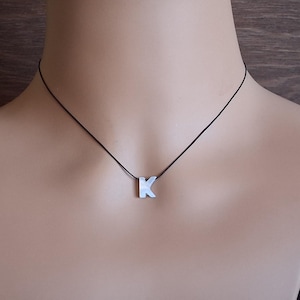 May include: A silver-colored pendant necklace featuring the letter "K". The pendant is suspended from a thin, black cord. The necklace is displayed on a mannequin.