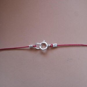 May include: A thin red cord necklace featuring a silver-toned spring ring clasp. The cord is secured at both ends by small silver-toned metal crimps that connect to the circular clasp mechanism, creating a simple and functional jewelry closure.