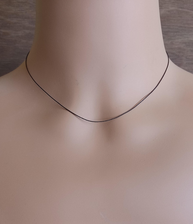 May include: A thin, dark brown, twisted wire necklace that rests closely against the neck. The necklace features a delicate, spiraled texture throughout its length, creating a subtle and refined appearance that sits smoothly around the base of the throat.
