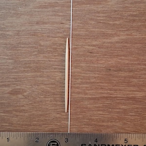 May include: A light-colored wooden toothpick positioned vertically next to a metal ruler. The toothpick measures approximately 2.5 inches in length, as indicated by the markings on the ruler visible at the bottom of the image.