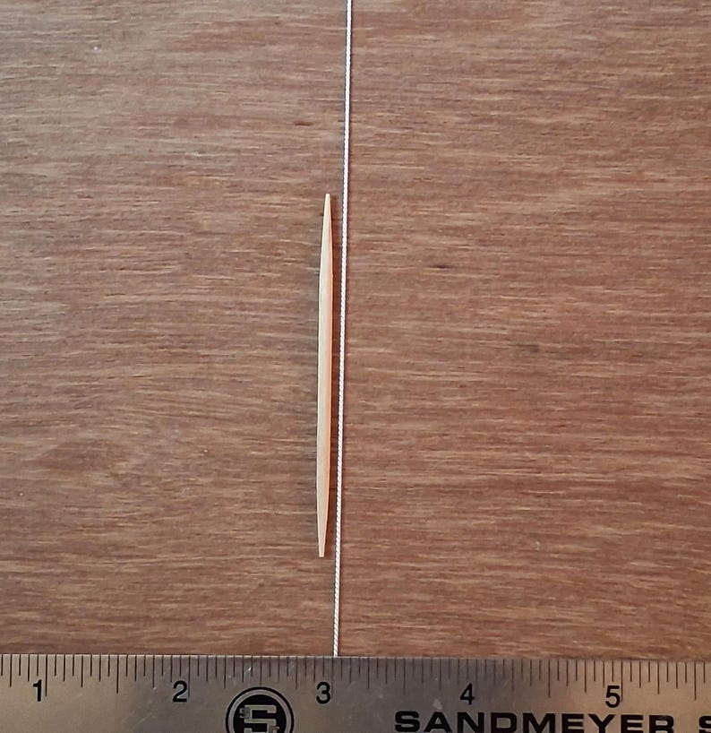 May include: A light-colored wooden toothpick positioned vertically next to a metal ruler. The toothpick measures approximately 2.5 inches in length, as indicated by the markings on the ruler visible at the bottom of the image.