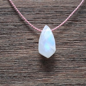 May include: A faceted, teardrop-shaped moonstone pendant with a translucent, milky white appearance and subtle blue iridescence, suspended from a thin, twisted pink cord that forms a necklace.