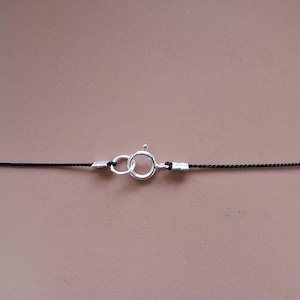 May include: A thin black cord necklace featuring a silver-toned spring ring clasp. The cord is secured at both ends with small silver-toned metal crimp beads that connect to the circular clasp mechanism, creating a simple and functional jewelry piece.