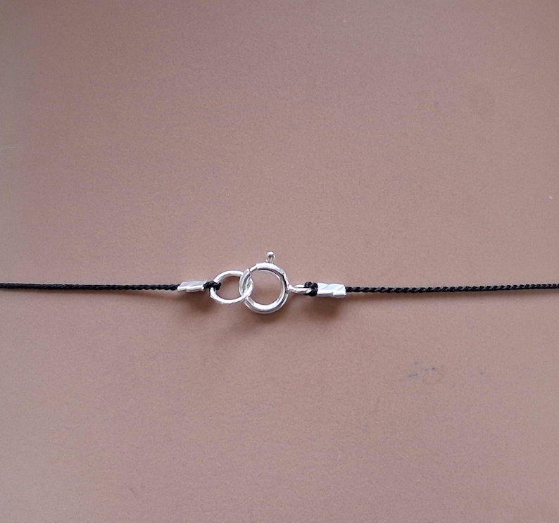 May include: A thin black cord necklace featuring a silver-toned spring ring clasp. The cord is secured at both ends with small silver-toned metal crimp beads that connect to the circular clasp mechanism, creating a simple and functional jewelry piece.
