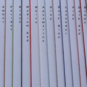 May include: A collection of fifteen vertical twisted cords in various colors, each labeled with text including Tan, Olive, Purple, Coral, Amber, Peridot, Black, Medium Red, White, Aquamarine, Gray, Dark Blue, Lavender, Pink, Dark Red, Brown, Gold, and Blue.