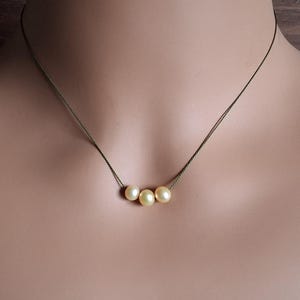 May include: A delicate necklace featuring three round, cream-colored pearls suspended on a thin, dark-toned cord. The pearls are arranged horizontally in the center of the cord, which rests against the skin, creating a simple and understated look.